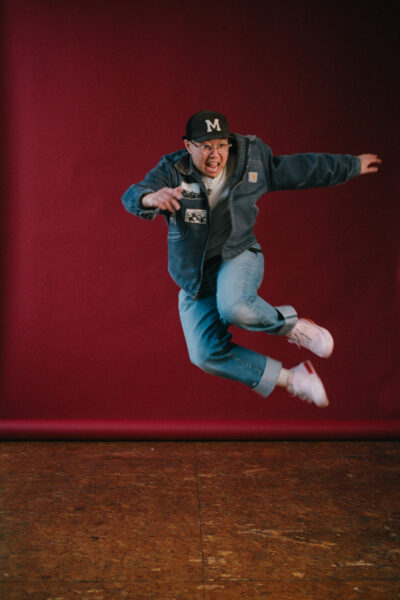A person wearing a cap, glasses, denim jacket, and jeans jumps energetically in front of a dark red backdrop.