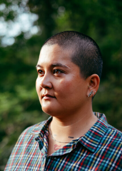 A person with a shaved head and multiple ear piercings, wearing a plaid shirt, stands outdoors with greenery in the background.