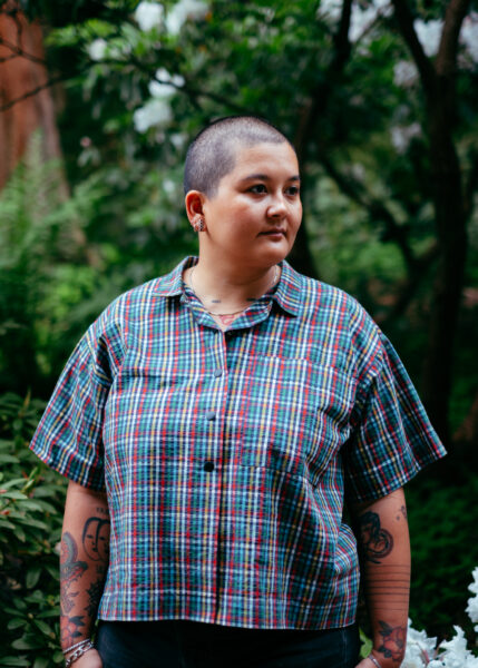 A person with a shaved head wearing a plaid shirt stands outdoors among green plants and trees, looking slightly to the side.