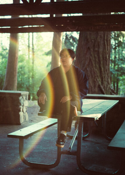 A person sits on a picnic table in a wooded outdoor shelter with sunlight creating rainbow lens flares across the image.