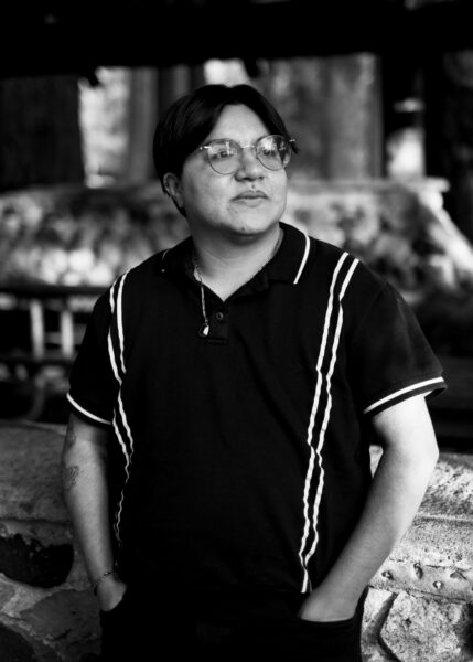 A person wearing glasses and a dark polo shirt stands outdoors with hands in pockets, looking off to the side. The image is in black and white.