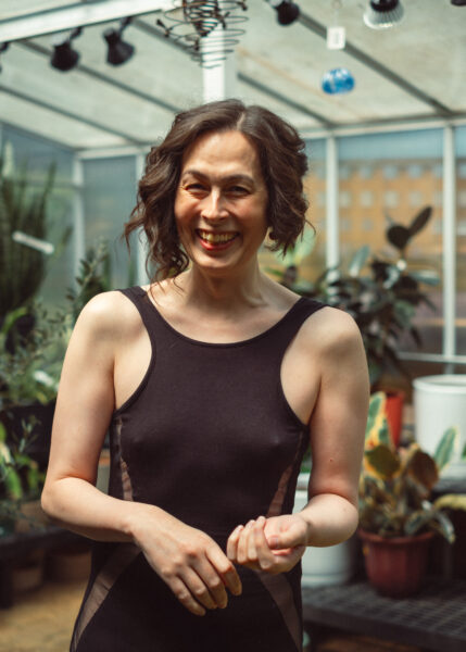 A woman with curly brown hair wearing a sleeveless black dress stands smiling in a greenhouse with various plants in the background.