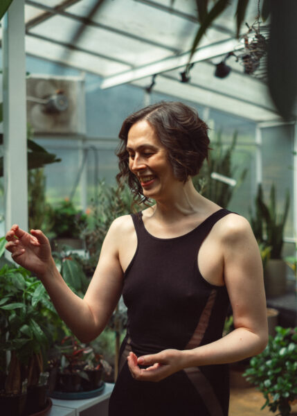 A woman in a sleeveless black dress stands smiling in a greenhouse surrounded by various green plants.