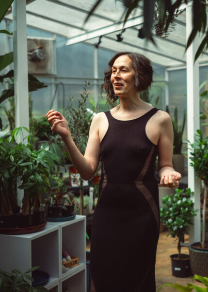 Person in a black sleeveless dress stands among potted plants in a greenhouse, gesturing with one hand and smiling.