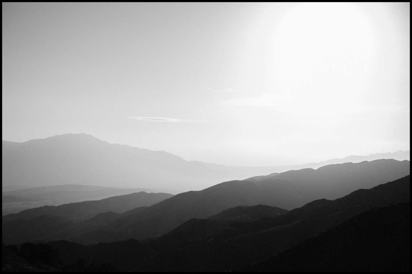 Black and white image of rolling mountain ridges under a bright sun with a hazy sky in the background.