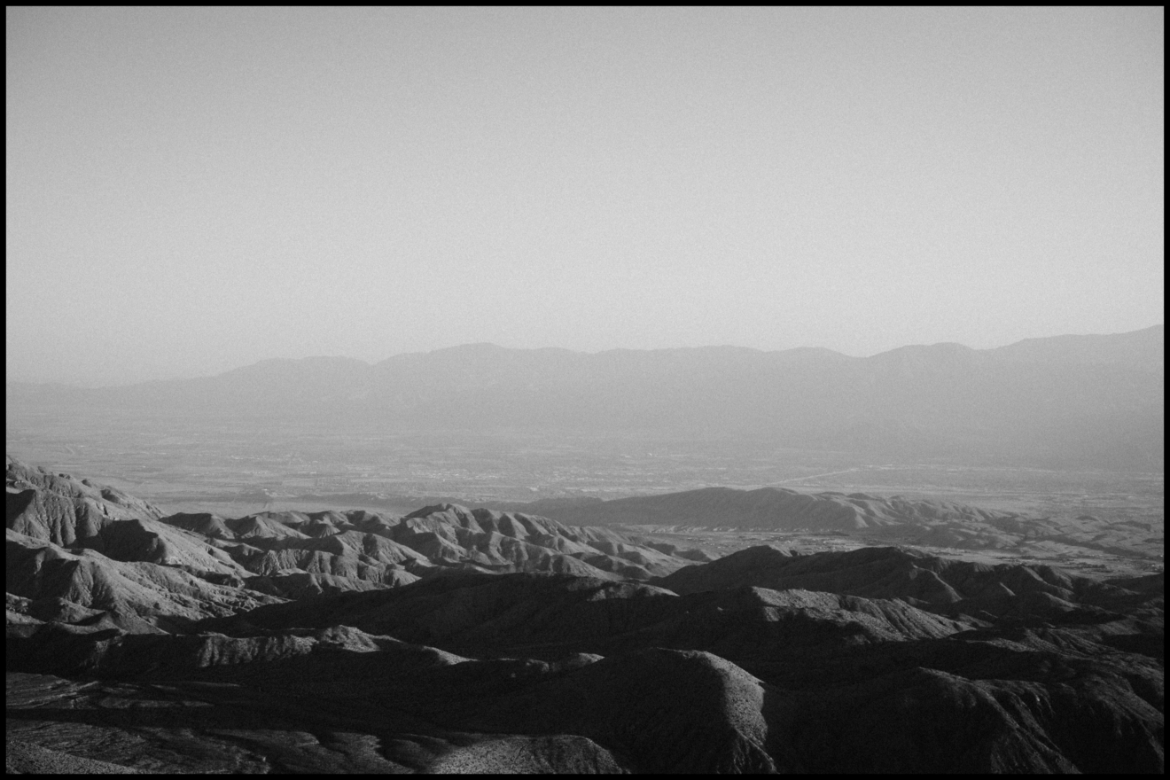Black-and-white photo of a rugged mountain landscape with ridges in the foreground and distant mountains under a hazy sky.