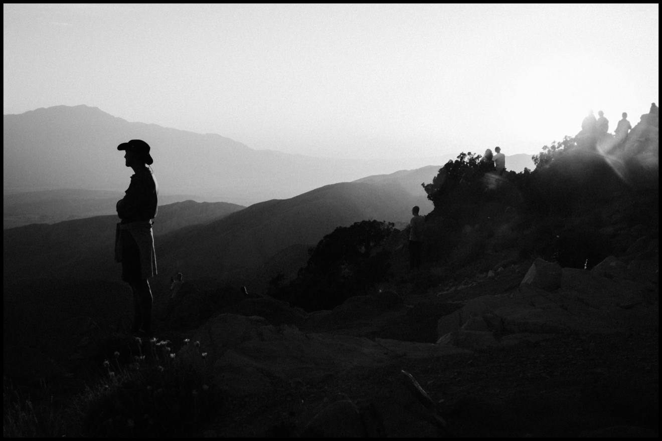 A person in a hat stands on rocky terrain at sunset, with other people and mountains visible in the background.