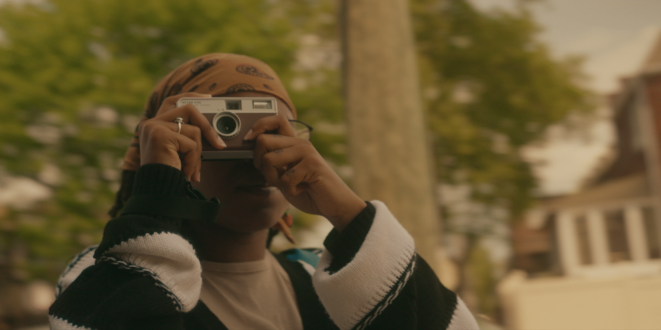Person wearing a headscarf and striped sweater holds a camera up to their face, taking a photo outdoors with blurred trees and houses in the background.