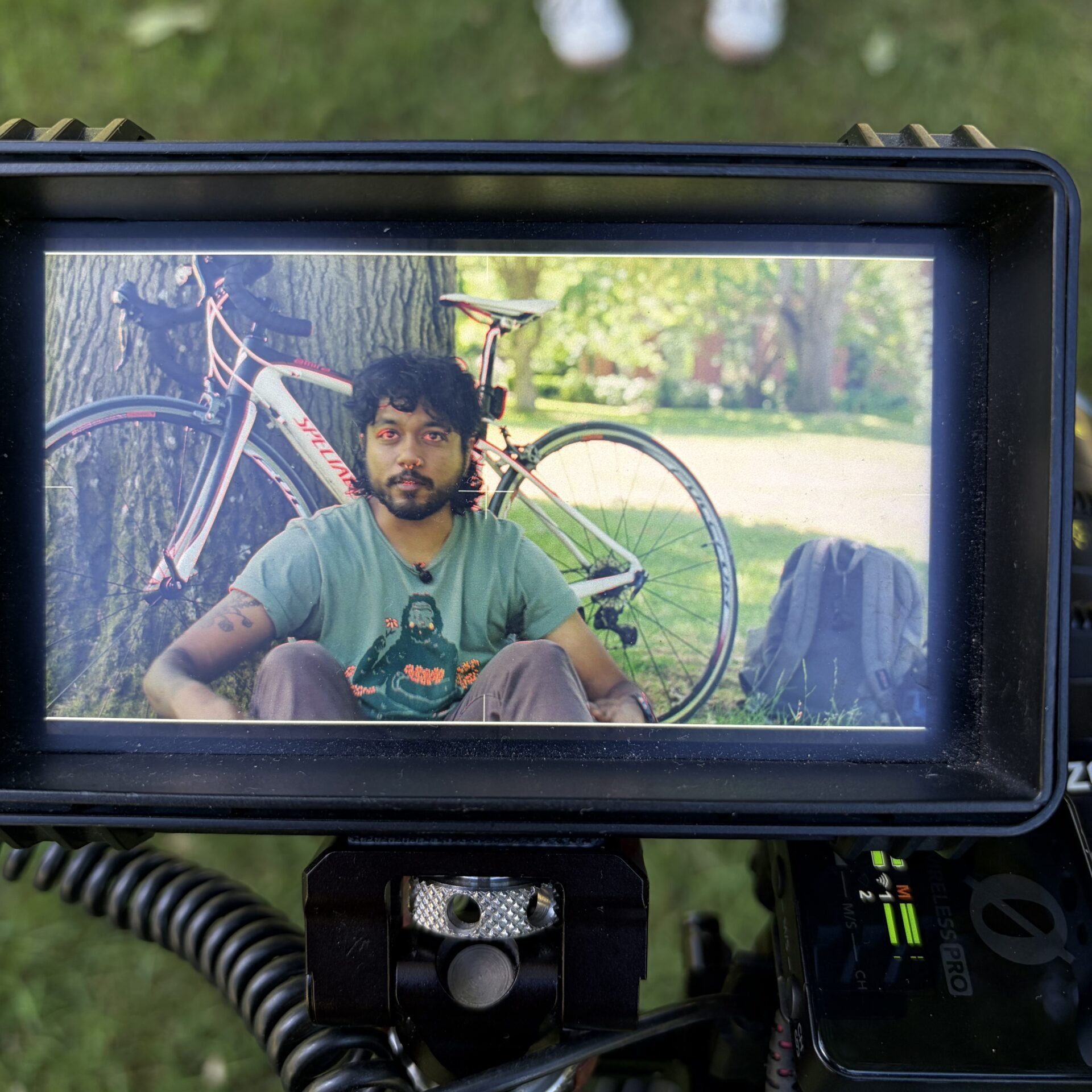 A camera monitor displays a person sitting on grass with a bicycle and backpack behind them; the scene is being filmed outdoors.