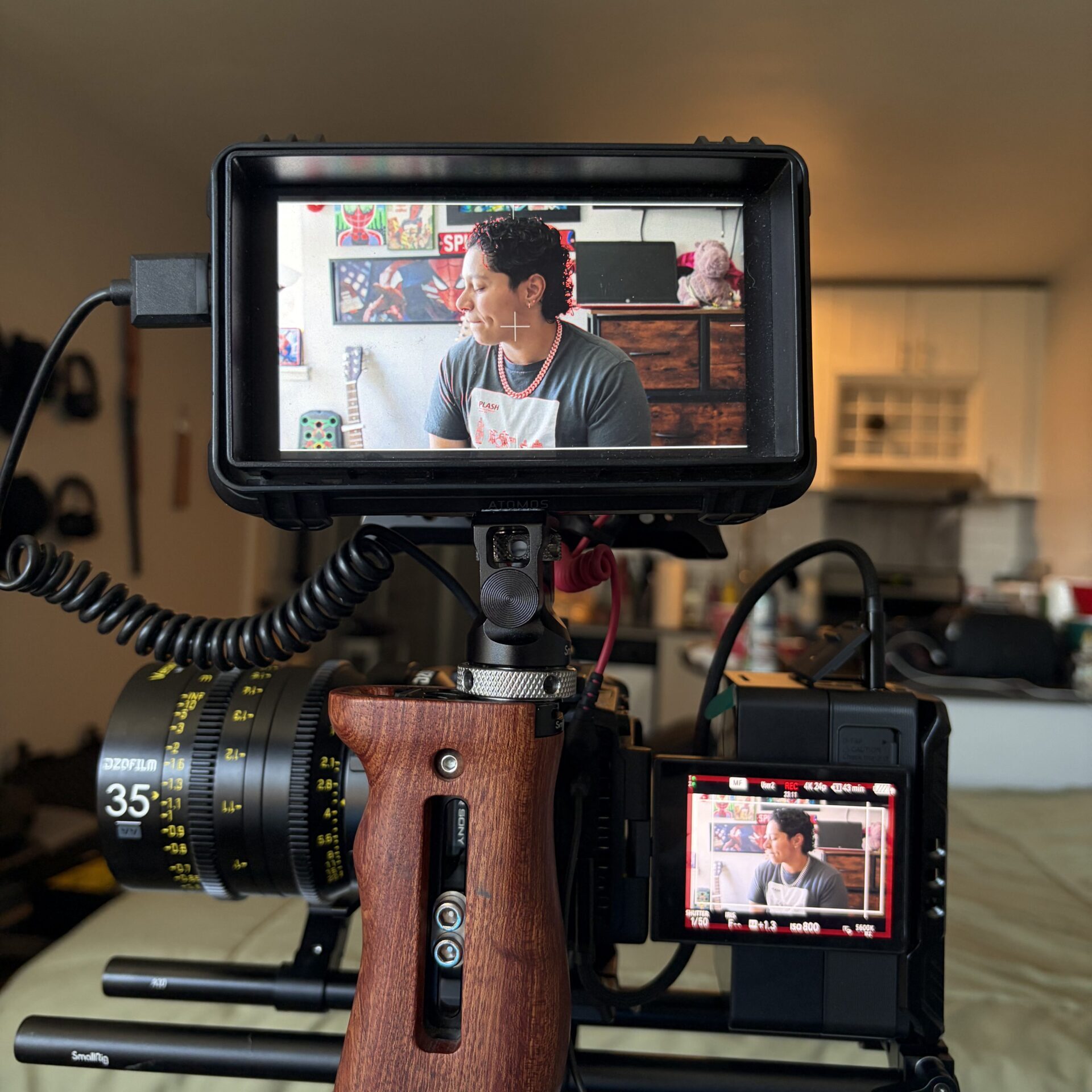 A video camera with a wooden grip films a person sitting in a bedroom, shown on the camera's screen display with kitchen and decorations visible in the background.