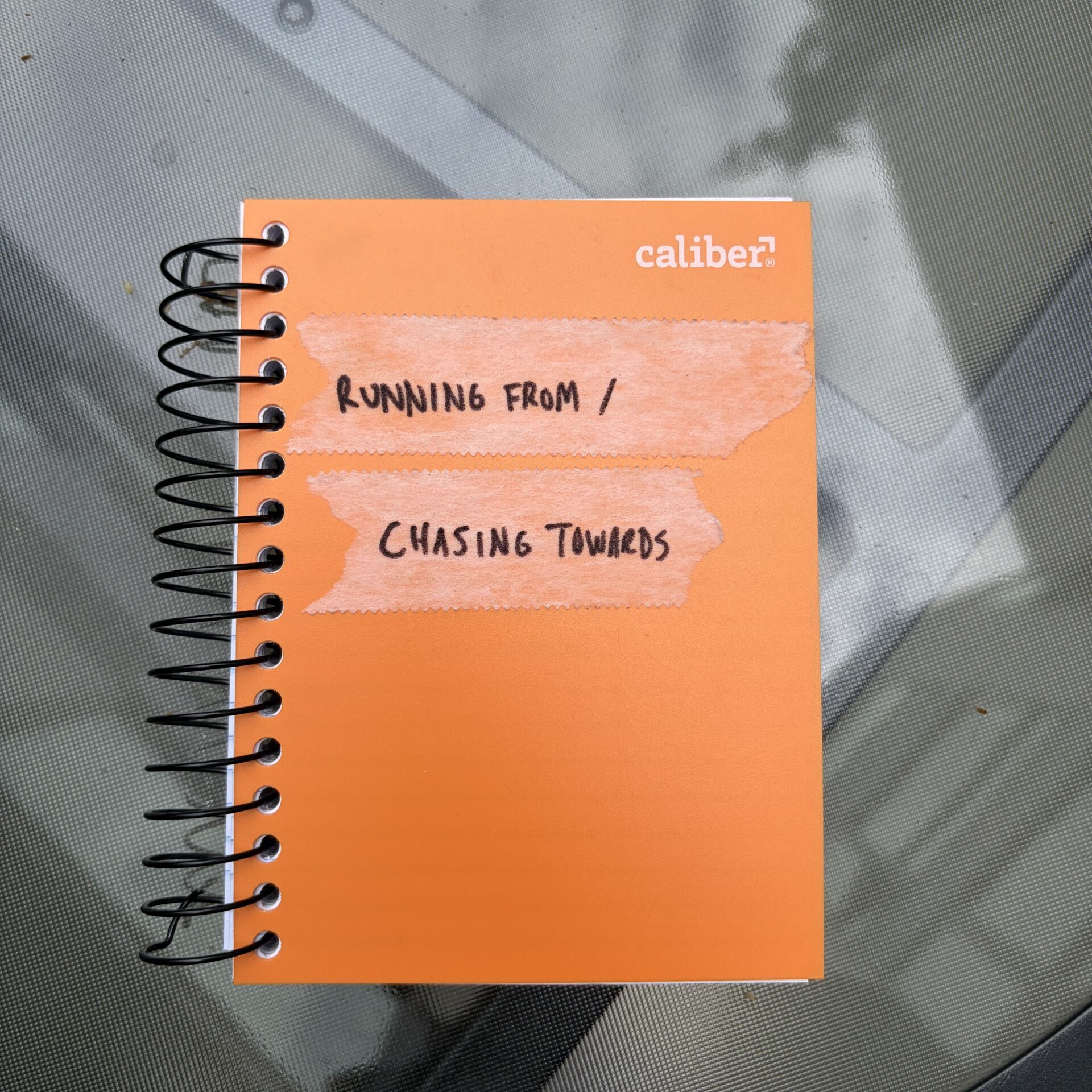 An orange spiral notebook with handwritten text on tape strips reads "RUNNING FROM / CHASING TOWARDS," lying on a glass table.