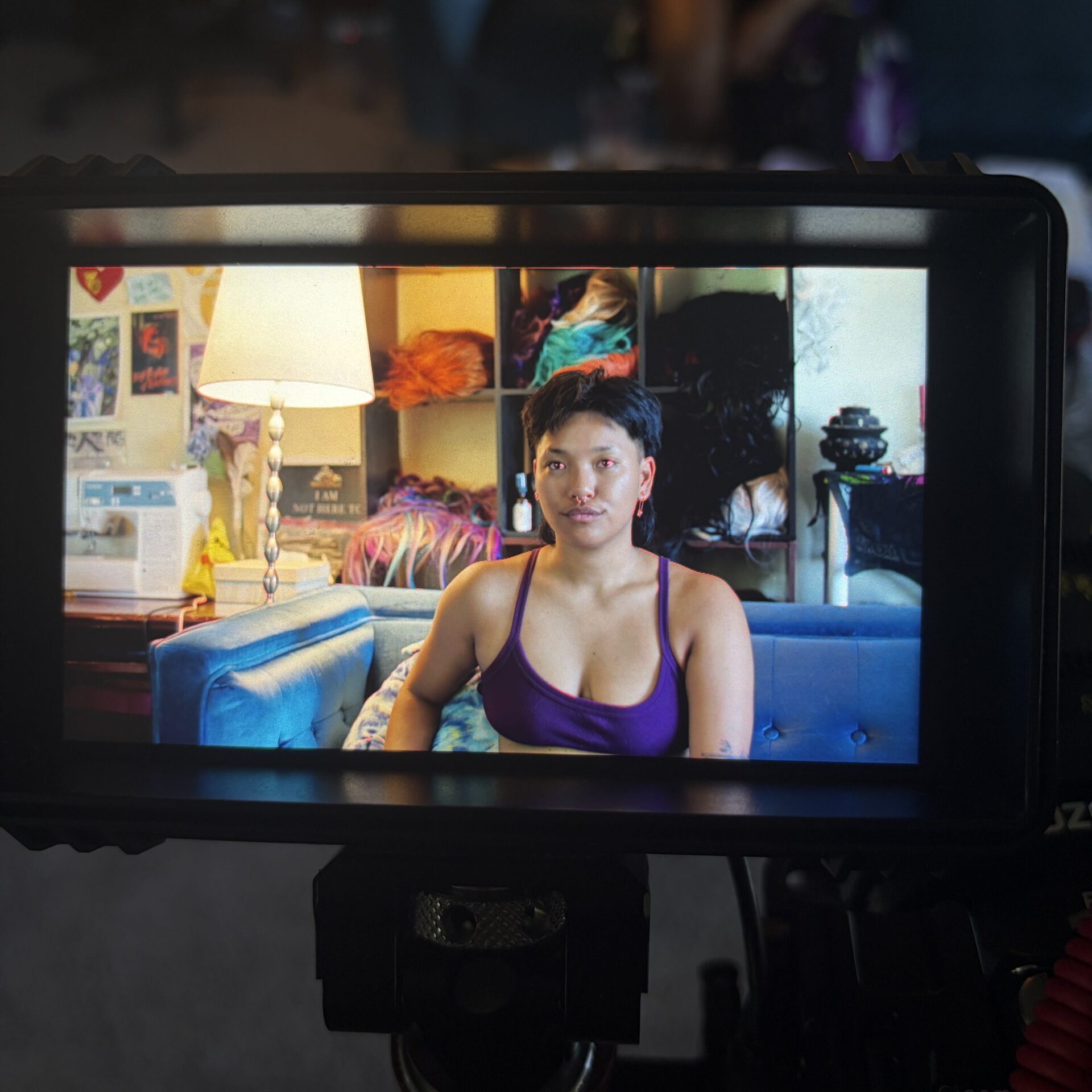 A person sits on a blue couch in a colorful room, viewed through a camera monitor, with a lamp, shelves, and sewing machine in the background.