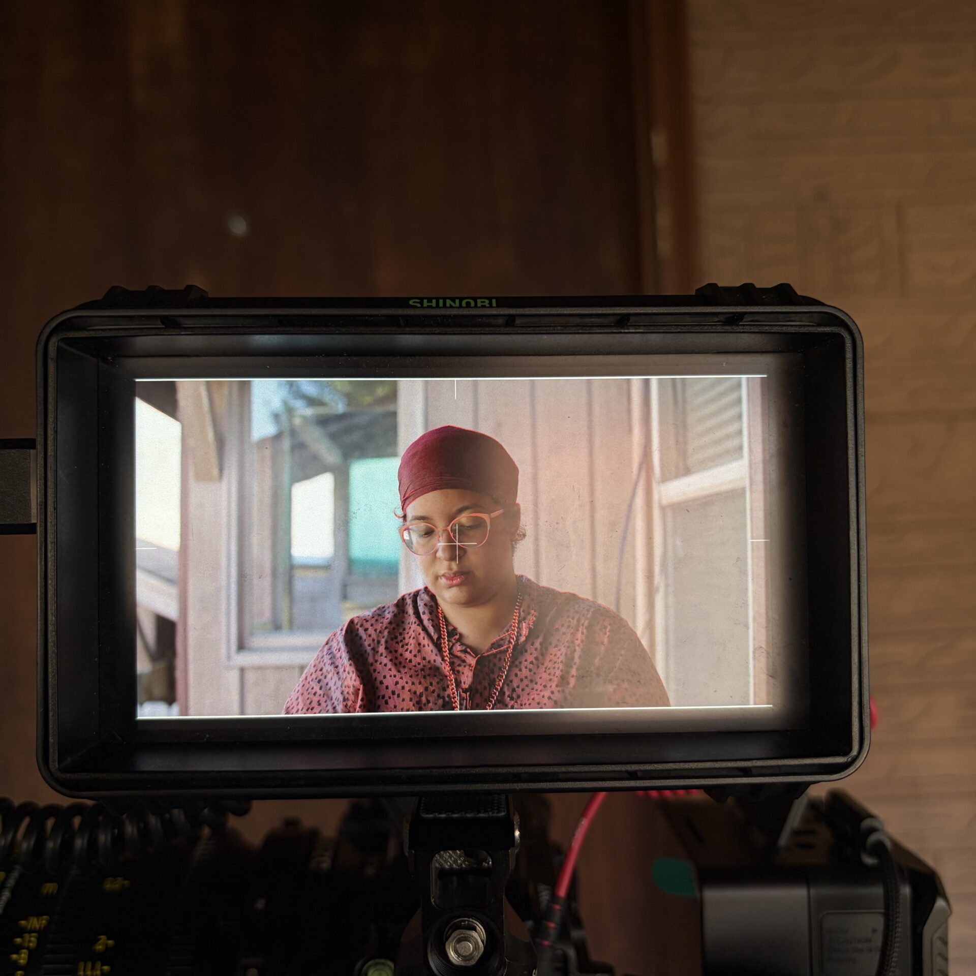 A camera monitor displays a person wearing glasses and a red head covering, seated indoors, with filming equipment visible in the foreground.