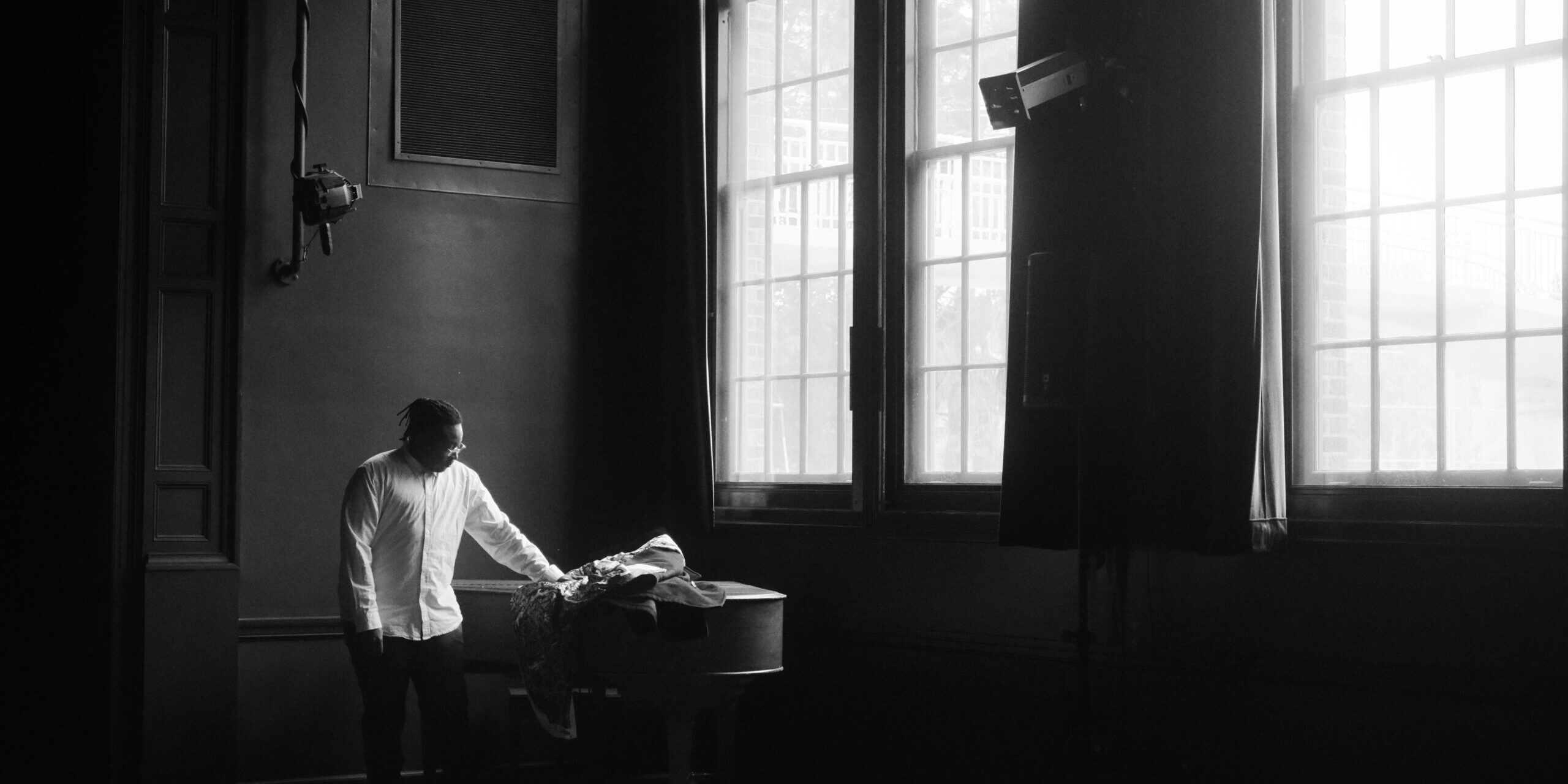 A person in a white shirt stands by a grand piano in a dimly lit room with large windows and wooden floors.